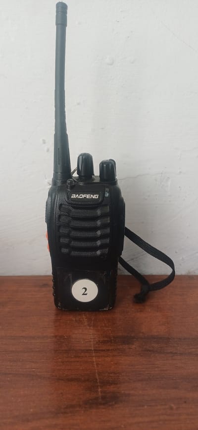 A black Baofeng walkie-talkie is positioned centrally on a wooden table. The walkie-talkie has a tall, thin antenna extending upwards. Two black knobs are visible on the top of the device. The brand name "BAOFENG" is printed in white on the front. A black lanyard is attached to the side of the walkie-talkie and is tied in a knot on the table. A white circular sticker with the number "2" in black is adhered to the bottom front of the walkie-talkie. The walkie-talkie is placed in front of a textured, off-white wall. The wooden table surface exhibits visible grain and some scratches. The scene appears to be indoors, and the lighting suggests it is daytime. There are no people or animals present in the image. Based on the information provided, this scene is not located in the city of Imeni Voroshilova, Kazakhstan.
