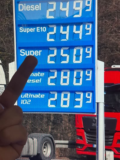 A close-up, slightly blurred image depicts a blue digital fuel price display at a gas station in Vellmar, Germany, observed during daylight hours. A light-skinned human hand with a visible index finger is positioned in the lower-left quadrant, pointing towards the "Super" fuel line displaying a price of 2.50⁹.
The digital sign, rendered in white text and segment numbers against a blue background, lists the following fuel types and prices in Euros per liter:
- Diesel: 2.49⁹
- Super E10: 2.44⁹
- Super: 2.50⁹
- Partially obscured text, likely "Ultimate Diesel" or similar, with a price of 2.80⁹.
- Ultimate 102: 2.83⁹
In the background, out of focus, a substantial portion of a red semi-truck is visible to the right of the sign, including its front grille, headlights, and a German license plate holder. Below the sign, a large, dark gray fender and a wheel, possibly belonging to the same or another heavy vehicle, are present. The far background shows defoliated trees or dense bushes, indicating an outdoor, possibly natural, setting beyond the gas station.