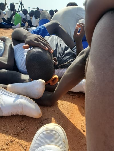 In Barakin Rafin Gora, Nigeria, a group of young men are gathered outdoors on a reddish-brown dirt surface, likely a field. The central focus is on one individual lying down, his head resting on the ground, with his hand placed on his forehead. He is wearing a white t-shirt and blue shorts. Another person's legs and arm are visible in the foreground, obscuring part of the central figure. Several other individuals are seated and lying down in the background, some wearing white shirts and blue shorts, and one is holding a green bag. The environment appears to be a sunny day, with a shadow cast across the ground. The scene suggests a break or rest period for the group.