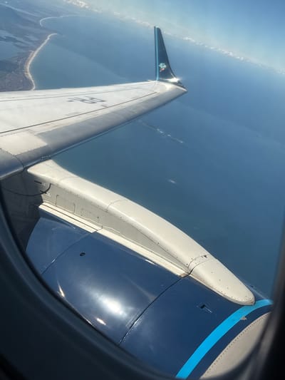 An aerial photograph captures the view from an aircraft window, primarily featuring the right wing and engine against a backdrop of ocean and coastline. The foreground is dominated by the aircraft's dark blue engine cowling, partially visible in the lower left and right, displaying a light blue horizontal stripe near its rear and reflecting bright sunlight. Above the engine, a light grey pylon connects it to the wing, exhibiting numerous rivets, panel seams, and stenciled markings reading "1-3" and "4-F". The main wing surface, also light grey, shows prominent black stenciling "H-25" on its upper side, along with visible panel lines and minor surface dirt or streaking. The wing terminates in a dark blue winglet, featuring a light blue vertical stripe on its trailing edge and a multi-colored, pixelated emblem on its outer surface, which also reflects sunlight.
Below the aircraft, a vast expanse of deep blue ocean covers the midground, appearing calm with subtle surface variations. Along the left side of the frame, a distinct coastline is visible, characterized by a light sandy beach forming a prominent curve. Inland from the beach, green vegetation is discernable, with what appears to be a narrow strip of land separating the ocean from a larger, calmer body of water further inland, suggesting an estuary or lagoon system. The sky above is a light blue, with faint, linear contrails or wispy clouds visible in the upper right background. The scene is brightly illuminated by direct sunlight. The geographical features suggest a coastal region in Brazil, specifically in an unknown city.