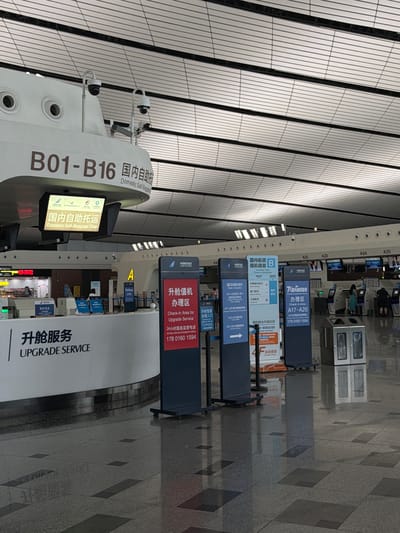 The image shows an airport check-in area with signs in Chinese and English. The signs indicate "Domestic Self-Baggage Drop" and "Upgrade Service." There are several check-in counters and computer terminals visible. People are seen standing at the counters and interacting with the staff. The environment appears to be a modern airport terminal with a tiled floor and a suspended ceiling with lights. The location is identified as Guangyang District, China.