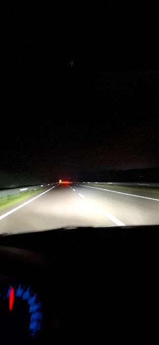 The image presents a nighttime, first-person view from within a moving vehicle on a multi-lane highway near Majmaa Tolba, Morocco. The road ahead is illuminated by the vehicle's headlights, highlighting distinct white lane markings, including solid lines on the shoulders and a dashed line in the immediate lane. Guardrails are visible along both sides of the roadway, reflecting the light. In the distance, several red lights, consistent with the taillights of other vehicles, are discernible. The sky above is dark. The lower portion of the frame shows the top edge of the vehicle's dashboard, and the lower-left corner reveals a segment of the instrument cluster with a red needle pointing at blue-illuminated markings. A faint green light is present in the dark upper-central area of the image. No individuals are visible.