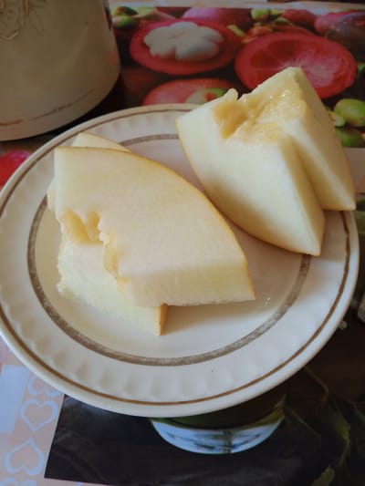 In Beşik-Jon, Kyrgyzstan, a close-up shot focuses on a plate holding slices of ripe melon. The melon slices, pale yellow and juicy, are arranged on a white plate with a gold rim. The foreground is dominated by these fruit slices, with a portion of a cream-colored pitcher or jug visible in the upper left corner. The background is indistinct but appears to be a surface with a colorful, possibly printed, design featuring fruits. The lighting is natural, casting a soft glow on the melon.