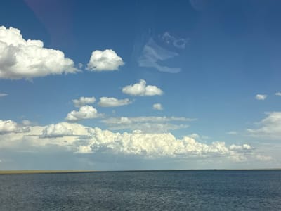 This media file captures a serene landscape view from a body of water, likely a lake or sea, under a vast blue sky dotted with fluffy white clouds. The foreground features the calm, rippling surface of the water, reflecting the sky above. A thin, flat strip of land, appearing as a yellowish-brown horizon line, separates the water from the sky.

The sky is a dominant element, filled with a variety of clouds. Some are large, cumulus formations, casting shadows and creating texture, while others are wispy and scattered. There's a faint, subtle rainbow visible near the center-left horizon, hinting at recent or ongoing precipitation. The lighting suggests it is daytime, likely late morning or afternoon, given the bright illumination of the clouds.

The scene evokes a sense of tranquility and openness. There are no people or man-made objects visible, emphasizing the natural beauty of the setting. The location context provided, Divnoe, Russia, suggests a potentially remote or rural environment. The weather appears pleasant, with a mix of sun and clouds, contributing to the peaceful atmosphere. There is no visible text in the image.