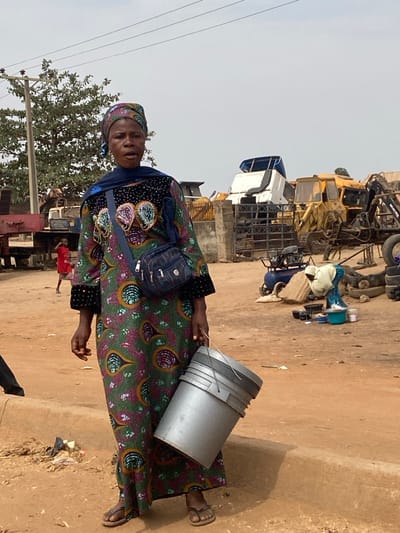 An adult female stands prominently in the foreground of an unpaved, reddish-brown dirt area. She is of African descent, facing the camera with a neutral, slightly open-mouthed expression. She wears a long, patterned dress with a dark top section featuring prominent embroidered designs in green, purple, and gold. A blue head covering drapes over her head and shoulders, and a dark blue shoulder bag with silver zippers is slung across her body. In her right hand, she carries two stacked grey plastic buckets by their wire handles. Her feet are clad in simple open-toed sandals.
In the mid-ground to the left, a child in a red garment is partially visible near large, dark-colored machinery. Overhead power lines and a power pole are present above a green-leafed tree.
The background on the right features a collection of industrial vehicles and heavy machinery, including yellow bulldozers, excavators, and white truck cabs, suggesting a salvage yard or equipment storage facility. A concrete barrier runs horizontally. Further to the right, another individual, dressed in light-colored clothing and a head covering, is bent over an arrangement of cooking or washing implements, including pots, pans, and a turquoise basin, on the dirt ground. A blue air compressor is also visible nearby. The sky above is overcast. The scene is located in the city of Lapai, Nigeria.