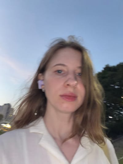 This is a close-up, waist-up, selfie-style photograph of a young Caucasian woman taken outdoors at dusk, likely in Uijeongbu-si, South Korea, given the presence of city lights and buildings visible in the background. The woman has long, light brown hair that is windswept and frames her face. She is wearing a white collared shirt, the fabric appearing textured. Her facial features are slightly blurred due to the nature of the selfie and low light conditions. She has blue eyes, a small nose, and pink lips. A notable detail is a large, light purple earring in her visible left ear, and a small silver hoop earring in her right ear. The background shows a hazy sky transitioning from light blue to a darker twilight hue, with the faint outline of buildings and some trees visible on the right side of the frame. The overall lighting suggests an urban environment at the end of the day.