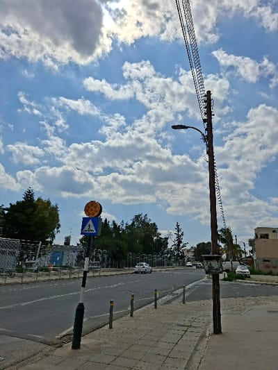 This image depicts a daytime street scene in Synoikismos Anthoupolis, Cyprus. The sky is partially cloudy with blue patches and scattered white cumulus clouds; the sun is visible in the upper left, partially obscured by a cloud. In the foreground right, a dark brown utility pole supports multiple black overhead electrical wires and a black street light fixture extending over the asphalt road. A metal box is attached to the pole at mid-height. The right side of the scene features a grey paved sidewalk, lined by several grey bollards with yellow reflective tops along the curb. Further left, a black and white striped pole supports a blue square pedestrian crossing sign with a white figure, topped by a circular amber reflector array. The two-lane asphalt road, marked with white dashed lines, stretches into the distance. A white car is observed moving away from the viewer in the left lane. In the background left, dense green trees and bushes are visible behind a grey metal fence or barrier. On the far right, behind the utility pole, a light-colored multi-story building is present, and a white vehicle is parked near it. No people are visible in the frame.