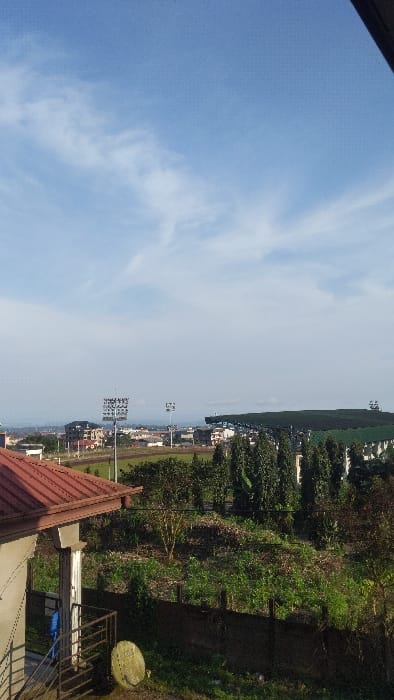 An elevated, static scene captures a daytime panorama in Buea, Cameroon, under a partially cloudy, light blue sky. In the immediate foreground, to the left, a portion of a building is visible, featuring a reddish-brown corrugated metal roof and a light-colored exterior wall. Metal stairs with a railing descend from the structure, where a blue fabric object hangs. A light-colored circular satellite dish rests on the ground near the stairs.
The midground consists of an overgrown, green vegetated area, interspersed with sparse bushes and some visible soil. A low, dark wall or fence partially recedes into this vegetation. Further back, a line of tall, dark green columnar trees stands.
The background is dominated by a large sports complex, identified as the Buea Omnisport Stadium. It features a green playing field, a prominent dark-colored grandstand with a roof, and several tall light towers equipped with multiple floodlights. Beyond the stadium, an urban landscape unfolds, displaying various residential and commercial buildings. Patches of exposed reddish-brown earth are discernible among the structures, suggesting development or open land. No individuals or active events are depicted in the scene.