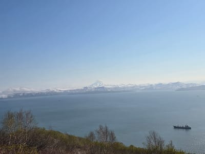 The image depicts a panoramic view of a large body of water, likely a bay or ocean, with a distant, snow-capped mountain range under a clear blue sky. In the foreground, a dense, green hillside is covered with dry brush and sparse, leafless trees, indicating a temperate climate. The water is calm and reflects the light blue of the sky, with subtle ripples visible on its surface. Several boats are present in the water: a small, dark vessel is positioned in the middle distance on the right, and two even smaller, indistinct vessels are further out towards the horizon. The distant mountains appear rugged and are partially obscured by a light haze, with patches of snow clinging to their peaks and slopes. The scene appears to be a natural landscape, possibly in or near the city of Krutoberegovoy, Russia, given the geographical features.