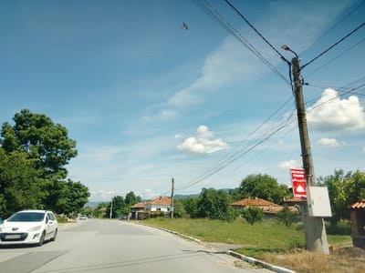 The image depicts a street scene in Mirovo, Bulgaria. A white Peugeot 308 station wagon is parked on the left side of a paved road. The car has a Bulgarian license plate reading "CO 5090 CM". On the right side of the road, a utility pole supports a street lamp and several power lines. A red sign with white lettering, indicating "RESTAURANT GRADINA" (Restaurant Garden) and "ОТВОРЕНО" (Open) with an image of a cloche, is attached to the pole. The surrounding environment consists of green trees, houses with terracotta roofs, and a cloudy blue sky with a bird in flight. The road appears to be in a rural or semi-rural area.