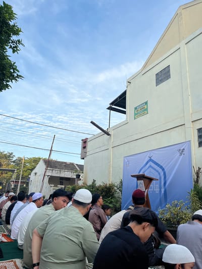 A gathering of Muslim men, most wearing white skullcaps, are praying outdoors on prayer mats. They are facing a makeshift podium with a blue banner behind it. The banner has text in Arabic. The background shows a building with a sign indicating "Rumah Di Jual". The overall location is determined to be in Sukabakti, Indonesia.