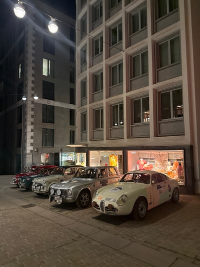 The image depicts a nighttime urban scene in Sankt Moritz, Switzerland, featuring a row of five vintage automobiles parked diagonally on a cobblestone-style street in front of two multi-story buildings.

The building on the right, occupying the majority of the frame, is a light-colored, multi-story structure with a grid facade of numerous windows. Its ground floor features brightly illuminated retail storefronts displaying clothing on mannequins, including items in yellow and orange hues. A reflection of "ZADAM" is visible on one shop window, and "LE CASINO ST. MORITZ" is visible on a decal on one of the parked cars.

To the left, a darker, taller building with distinctive light-colored quoining (decorative corner blocks) and a diamond-shaped motif on its facade is visible. Several of its windows are illuminated.

Five classic cars are parked, facing towards the viewer's left.
1.  The foremost car (right) is a cream-colored two-door sports coupe, possibly an Alfa Romeo, bearing the racing number "24" on its hood, doors, and side. Its license plate reads "H1267BBJ." It has a single auxiliary driving light mounted centrally on the front bumper.
2.  The second car is a silver-grey two-door sedan, consistent with an Alfa Romeo Giulia, featuring rally decals, including a shamrock and "LE CASINO ST. MORITZ" on its door. It has two auxiliary lights mounted on its front bumper and a license plate starting with "D-AV."
3.  The third vehicle is a light beige/white small sedan with rally markings.
4.  The fourth vehicle is a dark green small sedan, also with rally markings.
5.  The fifth vehicle, partially visible on the far left, is a red classic car with chrome bumper details.

Overhead streetlights, suspended by wires, illuminate the scene, casting reflections on the cars and facades. No individuals are visible. The vehicles are stationary.