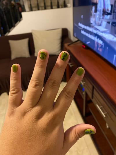 A hand with green nail polish and black designs is displayed in an interior setting. The thumb has two black circles, the index finger has "ГОЛБ" written on it, and the middle finger has a black smiley face. The ring and pinky fingers are painted only with green nail polish. In the background, a television screen is visible with what appears to be a medical scene displayed on it. A brown couch with white pillows and a brown shelf are also present. The location is possibly in La Aurora, Ecuador.