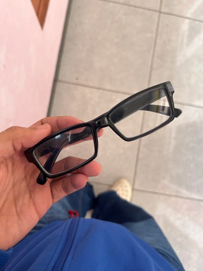 In Meteseh, Indonesia, a person is holding a pair of black-rimmed glasses with clear lenses. The glasses are positioned in the foreground, held by a hand with visible skin texture and knuckles. The background features a tiled floor and a blurred pink surface. The person is wearing blue clothing, including a visible zipper. A beige shoe is also visible in the lower right corner of the image.