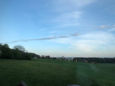The image depicts a landscape view in Kehrsatz, Switzerland, characterized by a green, grassy field taking up most of the foreground. In the middle ground, there is a scattered collection of houses and buildings including a water tower. Patches of trees and forest flank the sides. The sky above is light blue with some wispy, elongated clouds. The overall scene suggests a rural or suburban setting in the Swiss landscape.