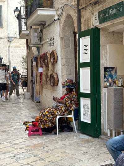 This image captures a vibrant street scene in a historic, stone-built town, likely Polignano a Mare, Italy. The paved street, made of light-colored stone, is lined with traditional buildings and features an arched passageway in the background.
In the mid-ground, a woman wearing a striking, multi-colored patterned dress and a matching headwrap is seated on a white plastic chair. She appears to be tending to a stall or display. Adjacent to her, a small pink stool is visible. Her display features several round, woven baskets hung on the stone wall, beneath which bundles of what appear to be straw or dried natural materials are arranged, possibly indicating a craft or local goods shop.
Behind the woman, a distinctive green shopfront stands out, featuring white panels. Visible text on the signage includes "Arti & Profumi" (Arts & Perfumes) and "DISTILLIAMO EMOZIONI" (We distill emotions), along with the website "WWW.ARTIEPROFUMIPOLIGNANO.IT". Another panel prominently displays the Italian quote "PERDUTO È IL TEMPO VISSUTO SENZA PROFUMO" (Lost is the time lived without perfume), reinforcing the theme of fragrances. The word "ESSENZE" (Essences) is also visible. Above the green shopfront, a poster for "MONTALE" perfumes is displayed on a table.
Further down the street, several people are seen walking, suggesting a pedestrian area during daytime. The scene is brightly lit, indicating clear weather. The setting conveys a lively, cultural atmosphere characteristic of an old European town, blending local craftsmanship with commerce.