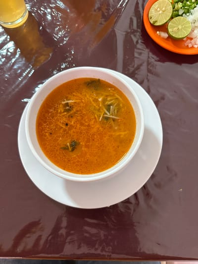 A close-up, overhead shot captures a bowl of soup on a glossy, dark brown table, presumably in Tizapan, Mexico. The soup, a vibrant orange broth, is filled with thin noodles and small green herbs. It sits in a white bowl, which rests on a white saucer. To the upper right of the bowl, an orange plate holds two lime wedges, chopped white onions, and green jalapeños, suggesting a side accompaniment. A partial view of a glass filled with a pale yellow liquid is visible in the upper left corner. The table surface is covered with a clear, crinkled plastic tablecloth that reflects the light, creating a dynamic, almost liquid appearance. The overall scene suggests a casual dining setting, likely a restaurant.