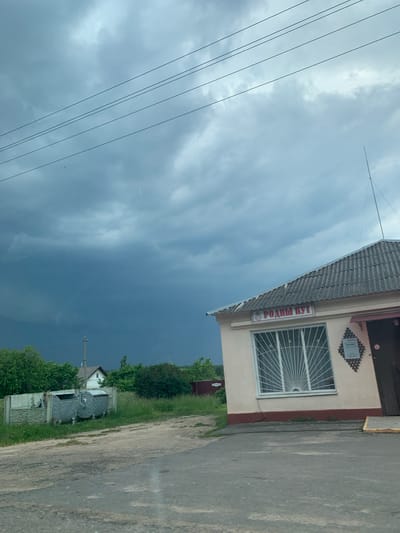 The image shows a roadside scene with a small building and a stormy sky. The building, which appears to be a shop or kiosk, has a light-colored facade with a darker trim around the base and a corrugated roof. A sign above the window reads "РОДНЫ КУТ" in red lettering. The window is covered with a decorative white metal grate. To the left of the building, there are two large metal trash bins. In the background, there are trees, a fence, and a house. The sky is dark and overcast with visible storm clouds. The overall impression is a rural or semi-rural setting in Слабада-Кучынскі сельскі Савет, Belarus, under threatening weather conditions.