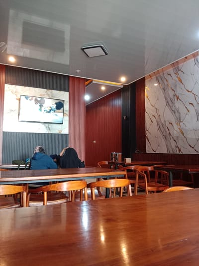 The image depicts an indoor setting, likely a restaurant or cafe, in Pandean, Indonesia. The foreground features a large, polished wooden table, with several wooden chairs around it.
Two individuals are seated at a table further back, engaged in watching a television screen mounted on a wall. One person is wearing a blue hooded jacket and the other is in black attire. The wall behind them is decorated with a combination of grey striped panels and a marble-effect section.
The overall environment has a modern aesthetic, with a glossy grey ceiling and recessed lighting. Another wall with a similar marble design is visible to the right. Various wooden tables and chairs fill the scene.