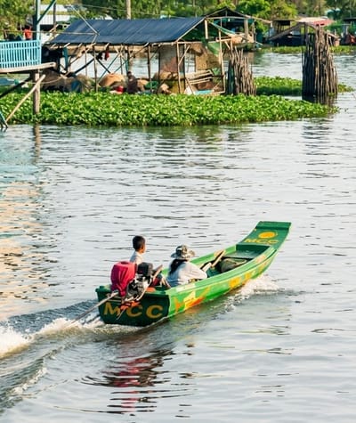 In Soutr Nikom, Cambodia, on a body of water, two individuals are in a narrow, green motorized boat. The person at the rear is steering the boat, while the person in front is looking forward. The boat is creating a wake as it moves. In the background, there is a stilted structure on a bank covered in lush green vegetation. The sky is a light blue, suggesting daytime. The overall scene depicts a tranquil, rural riverine environment.