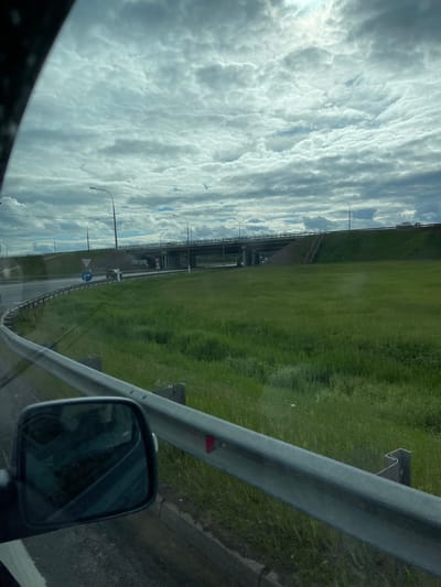 The image shows a view from inside a vehicle, likely a car, traveling on a highway near the city of Starokurovo, Russia. The scene is viewed through the side window, with the side mirror partially visible. A grey metal guardrail runs along the right side of the road. Beyond the guardrail, a grassy slope rises towards what appears to be another road or overpass. Street lights are visible on the left side of the road. The sky is filled with layered clouds. The road surface is visible, and some road signs are discernible in the distance.