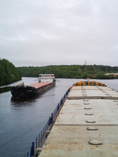 The image depicts a river scene in Depo, Russia, under an overcast sky. Two barges are visible. The first barge is black and carries a reddish-brown load. The name "Volgodonsk" is written in Cyrillic letters on its side. The second barge, positioned closer to the viewer, has a gray deck with a series of oval hatches and yellow equipment at its bow. Both barges are on a river bordered by green trees on either side. A power line is visible in the distance.