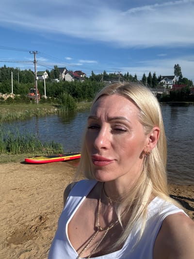 A medium shot shows a blonde woman in a white top posing for a selfie outdoors. She has light skin, blonde hair, and is wearing jewelry. Behind her is a body of water, a sandy shore with a red and yellow paddleboard, and lush green foliage. In the background, several houses, trees, and utility poles are visible under a blue sky with scattered clouds. An orange tractor is also visible in the distance. The scene appears to be a rural or suburban area.