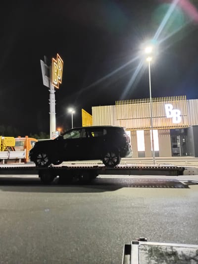 A black SUV is being transported on a flatbed tow truck at night in Drahelčice, Czechia. The SUV is positioned over the rear of the tow truck. In the background, a building with a modern facade featuring vertical wooden slats and large illuminated "BB" signage is visible. A tall pole with a double "BB" illuminated sign stands to the left of the building. A bright street light illuminates the scene, casting a strong glare. The ground appears to be asphalt, suggesting a road or parking lot. The sky is dark, indicating nighttime.