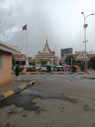 The image depicts a cloudy day in Bavet Quarter, Cambodia. In the foreground, a wet asphalt area with puddles and tire tracks is visible, bordered by a yellow and gray curb. To the left, a light orange building with ornate white trim is partially in frame. In the midground, a large flagpole with the Cambodian flag flies against the sky. Behind the flagpole are several buildings with traditional Cambodian architecture, featuring tiered, orange-tiled roofs and decorative spires. A line of vehicles, including white SUVs and vans, are parked in front of these buildings. Several people are seated under red and blue umbrellas, appearing to be vendors or customers at small stalls. In the background, a tall, unfinished high-rise building stands out against the overcast sky. A street lamp with multiple lights and a solar panel is positioned on the right side of the frame. The overall scene suggests a public area, possibly a market or a transportation hub, given the presence of vehicles, stalls, and the national flag.