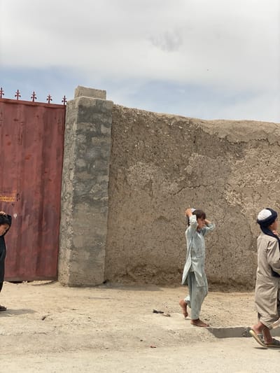 The image depicts a street scene in Panjwai, Afghanistan. A weathered, textured wall of mud and concrete stretches across the background, with a red metal gate on the left side. Three children are visible walking on the dirt and concrete sidewalk. The child in the middle is barefoot, while the child on the right is wearing sandals. The children are dressed in traditional clothing. The sky is partly cloudy.
