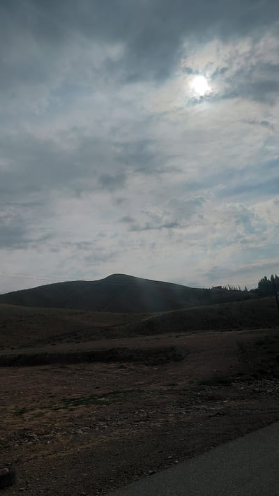 The image captures a dramatic landscape under a cloudy sky, with the sun partially obscured by the clouds, suggesting either early morning or late afternoon. The scene is dominated by a steep, barren hillside to the right, sparsely covered with rocks and dry vegetation. A narrow, dark road runs along the edge of the hillside, its surface appearing to be asphalt or compacted gravel.

To the left, the sky is filled with a mix of dark and light clouds, with the sun's bright rays piercing through, creating a dramatic and somewhat diffused lighting effect. The clouds are wispy in some areas and more solid and grey in others, hinting at dynamic weather conditions.

Along the crest of the hill, a line of dark, slender trees stands silhouetted against the sky. Below them, on the slope of the hill, a faint, almost imperceptible power line runs diagonally downwards, supported by distant, thin poles. The overall impression is one of a remote, rugged, and natural environment, possibly in a mountainous or hilly region, as indicated by the topography. The lack of any clear human figures or distinct structures, apart from the road and the power line, emphasizes the vastness and wildness of the location. The mood is somewhat solemn and awe-inspiring, due to the dramatic sky and the imposing natural features. The time of day is suggested by the diffused sunlight, likely not midday.