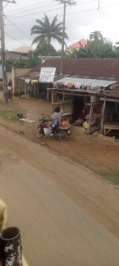 This image captures an outdoor scene in Ezinifite, Nigeria, under an overcast sky. A wide, reddish-brown dirt road occupies the foreground and midground.
In the midground, a person wearing a patterned top and shorts is seated on a red and black motorcycle, parked at the edge of the road, partially on a low concrete structure. A brown, cylindrical container is strapped to the motorcycle's rear rack. The individual's right arm is raised, hand open. Directly behind the motorcycle and to the right, a series of simple, open-front market stalls or shops with rusted corrugated metal roofs are visible. Colorful garments hang on lines beneath the eaves of these roofs. Inside one stall, a person is seated.
Further to the left, two additional figures are visible near a structure, one partially obscured in a long garment. Overhead utility poles and numerous power lines traverse the upper portion of the frame. In the background, dense green vegetation, including a prominent palm tree, partially obscures additional buildings, one featuring a distinct red roof. A white banner with black text, partially legible as "A.C.M.F. FEMALE...", is affixed above one of the market stalls. A dark, cylindrical object, likely part of the camera's immediate surroundings, is visible in the bottom-left corner of the frame.