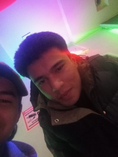 This image depicts two individuals in an indoor setting within Takhtakuprik, Uzbekistan. The main subject, a male, occupies the right half of the frame, positioned towards the camera with a neutral expression. He has short, dark, textured hair and is wearing a dark, possibly padded jacket with a lighter-colored collar or hood lining visible. A silver-toned metal eyelet is visible on the jacket's lapel area. The second individual is partially visible on the left, showing only the upper portion of their head and face, wearing a dark flat cap, and appearing out of focus. The background consists of light-colored walls illuminated by multiple colored light sources: a strong pink and purple hue on the upper left ceiling and wall, and a green glow on the upper right wall. A red light accentuates the right side of the main subject's head. A red and white sign with reversed Cyrillic text and an unidentifiable graphic is visible on the wall behind the subjects. A ceiling fixture is partially visible in the upper background.