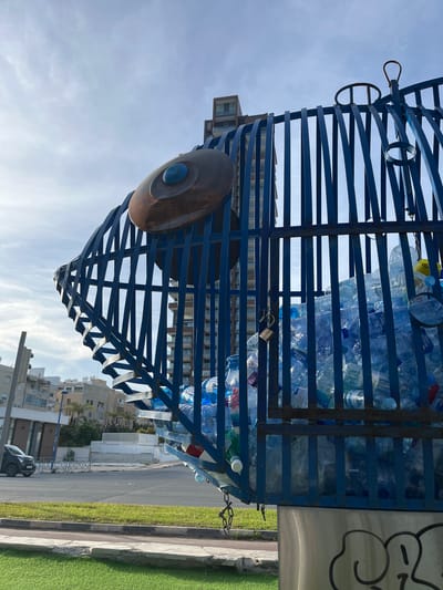 The image depicts a metal trash bin filled with used plastic water bottles, located in Germasogeia, Cyprus. The bin is constructed of blue-painted metal bars arranged in a cage-like structure. A brown, disc-shaped object with a blue center is attached to one side of the bin. The bin is positioned on the side of a street with buildings and greenery visible in the background. A sidewalk borders the street, with green artificial turf along one section. A parked car is visible in the distance. The sky is visible above the bin and buildings, with some clouds present.
