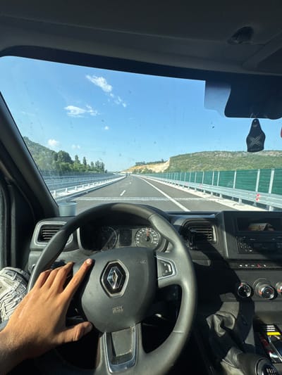 The image captures a first-person perspective from the driver's seat of a vehicle, facing forward through the windshield. The primary subjects are a person's hand on the steering wheel and the interior of a truck's cab. The scene is a highway, evidenced by the asphalt road with white lane markings and a protective barrier along the side. Green sound barriers line the highway on the left, with a green, hilly landscape beyond them. The sky is clear and blue, with a few scattered white clouds, suggesting fair weather.
The time of day appears to be daytime, likely late morning or afternoon, given the bright sunlight casting shadows inside the cab. The steering wheel is black and features the Renault logo in the center. The dashboard includes a speedometer, air vents, and various controls. A small black tag with a white "STYLE" logo hangs from the windshield. The person's hand, resting on the steering wheel, has a watch on the left wrist. Visible text includes "STYLE" on the hanging tag, "R N" on the steering wheel, and markings on the dashboard gauges, one of which appears to read "120." The activity is driving on a highway.
The location context provided is Peshterna, Bulgaria. While there are no explicit visual cues in the image that directly point to Bulgaria, the overall setting of a modern highway through a rural, hilly landscape is consistent with such a location. The image conveys a sense of travel and movement.