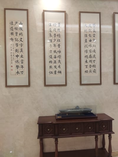 The image shows a wall with three framed calligraphic artworks and a wooden table with a model aircraft carrier. The artworks feature vertical columns of Chinese characters, written in black ink on a white background and framed with light brown wood. The characters appear to be a form of traditional Chinese calligraphy.

The leftmost artwork has smaller characters on the left margin, possibly a signature or date. The middle and rightmost artworks are similar in style. The calligraphic text itself is difficult to discern precisely without higher resolution or knowledge of the specific script and content.

Below the artworks, a dark brown wooden table with carved details and brass drawer pulls is visible. On top of the table, a model of a large aircraft carrier sits inside a clear display case. The model is grey and appears to be quite detailed.

The wall behind the artworks is tiled with light-colored, large square tiles. The lighting in the room appears to be from overhead, casting a slight shadow on the wall. The overall impression is one of a formal display area, possibly in a museum, gallery, or a well-decorated residence.

The question about the location being in Ürümqi, China, cannot be confirmed or denied from the visual information provided in the image. There are no landmarks, signs, or other contextual clues within the image that specifically identify it as being in Ürümqi.