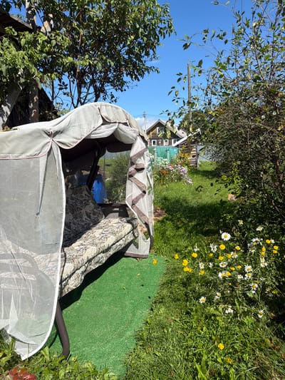 The image depicts a sunny outdoor scene in Yalguba, Russia, likely a backyard. In the foreground, a fabric-covered garden swing is set on a patch of artificial green turf. The swing is draped with a white mosquito net, and its cushions have a patterned upholstery. To the right of the swing, a cluster of white and yellow wildflowers grows in the grass.
In the background, several wooden houses with gabled roofs are visible, suggesting a rural or semi-rural residential area. A green fence separates the properties, and beyond that, trees and vegetation are abundant. The sky is a clear, bright blue. The overall impression is one of a quiet, summery day in a Russian dacha environment. No people are present in the image.
