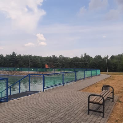 This image depicts an outdoor recreational area with a body of water and a paved walkway. In the foreground, a wooden bench with a metal frame sits on a gray paver surface adjacent to dry, yellowed grass. The walkway, also paved with gray pavers, extends to the left, bordering a fenced-off section of water. The fence consists of blue metal posts and green mesh. Further back, a row of dark green coniferous trees forms a dense backdrop. The sky above is blue with scattered white clouds. A tall wooden utility pole with wires is visible in the distance. The scene appears to be in or around Dubrivka, Ukraine, suggesting a park or a similar public amenity.