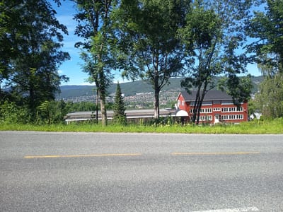 A road runs across the foreground, marked with a single yellow line. Beyond the road is a strip of green grass and vegetation, with several large trees framing the view. In the background, a large, red building with a dark roof is visible, featuring numerous white-framed windows and a white door. To the left of the red building, a long, lower building with a grey roof and many windows is partially obscured by trees. Further in the distance, a hillside covered in trees and a dense residential area is visible, with a clear blue sky overhead. The image appears to have been taken on a sunny day, likely in the late morning or early afternoon, given the bright sunlight and clear shadows. The setting suggests a suburban or semi-rural area, possibly a school or community center given the architecture of the buildings. No people are visible in the image, and there is no visible text. The overall impression is one of a peaceful, sunny day in a picturesque location.