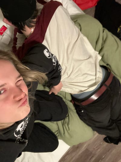 The image captures an overhead, slightly tilted view of two individuals lying down, likely on a bed. The perspective makes the person in the foreground appear inverted.

In the foreground, a person with blonde hair is visible, looking upwards towards the camera. Their eyes are partially open, and their lips are slightly parted. They are wearing a black long-sleeved top, possibly a hoodie, which features white graphics or symbols on the sleeve. Their left arm is bent, and a small part of their hand is visible near their head.

Adjacent to and slightly behind the first person, a second individual is lying down. This person is wearing a light-colored (cream or off-white) long-sleeved top with distinctive burgundy-colored lower sleeves or cuffs. They are also wearing black pants and a brown belt, which is visible around their waist. A dark, possibly black puffer-style jacket, is draped over their upper body. Their head is mostly obscured by the first person's arm and hair, but a glimpse of dark hair can be seen. A silver-colored bracelet or band is visible on their wrist.

The setting appears to be a bedroom or a cozy indoor space, characterized by a light green blanket or comforter covering a white sheet. A wooden floor is visible in the upper right portion of the background, suggesting the edge of the sleeping area.

Near the blonde-haired person's arm, on the white sheet, there is a small red object and a small white object, possibly a charger or a small electronic device. The overall activity suggests a relaxed moment, possibly a casual selfie or photograph taken while lounging. No discernible text is visible within the image.