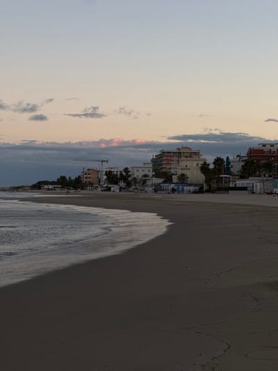 The image displays a coastal scene in Grottammare, Italy, observed at twilight. The foreground features a dark brown-grey sandy beach, bordered on the left by the sea with small breaking waves. The wet sand line delineates the water's edge. In the midground, the beach extends, transitioning to a strip of urban development in the background. This urban area comprises multi-story buildings, including structures with pale facades, one with a prominent red roof, and another with an exposed light-orange exterior. A large white construction crane is visible above the buildings in the mid-left. Palm trees are interspersed among the structures. Along the beach, in front of the buildings, numerous white and light blue individual beach cabins or changing huts are aligned. A small, distant red object, possibly a boat or rescue equipment, is situated on the far right of the beach. A few human figures are faintly discernible on the wet sand in the distant left-midground. The sky above is a gradient of pale blue, transitioning to light pink and orange hues near the horizon, accented by scattered gray and pink-illuminated clouds.