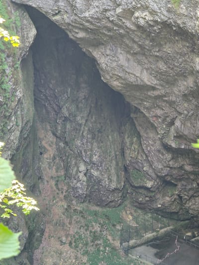The image shows the entrance of a cave with a large rocky overhang. The cave entrance is dark and appears deep. The surrounding rock faces are rough and textured, with patches of moss and vegetation. In the lower right portion of the image, there is a fenced-off area with a paved surface and some construction materials, suggesting some form of development or pathway near the cave. The cave is located in the city of Vilémovice, Czechia, likely a natural attraction or geological formation.