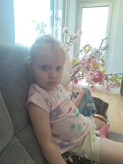 The media file depicts a young child lying on a sofa. The child has blonde hair and blue eyes and is wearing a light pink t-shirt with a floral pattern. Beside the child, there is a vase of artificial pink and white flowers, possibly cherry blossoms, positioned in front of a large window. The room appears to be indoors, with a sofa that has a textured grey fabric. The lighting suggests it might be daytime, with bright light coming from the window. In the background, a black bag and some other objects are partially visible, as well as what appears to be a wooden floor. The child's expression is neutral, and they are looking towards the camera. The scene is likely a home environment. There is no visible text in the image.
