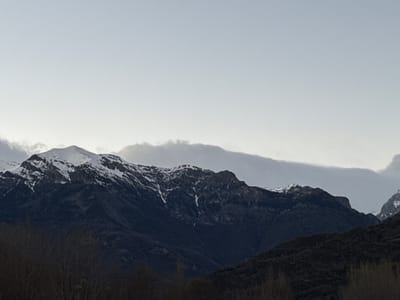 The image shows a mountain range partially covered in snow, likely taken near Castejón de Sos, Spain. The sky is overcast and gray, providing soft, diffused light. In the foreground, there are dark, leafless trees, indicating either a winter or early spring season. The mountains are rugged with visible rocky outcrops and patches of snow clinging to the higher elevations. The overall tone is muted and somber.