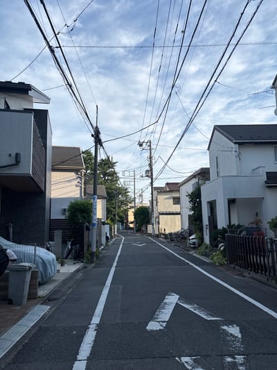 A straight-on, ground-level shot depicts a residential street in Suginami, Japan. The asphalt road is flanked by two-story homes on either side, the architecture a mix of modern and traditional Japanese styles. Overhead, a network of power lines crisscrosses the sky, obscuring a partly cloudy blue sky. A white directional arrow painted on the road points to the left. Street signs and typical Japanese street furniture (covered items, utility poles) line the roadside. No people are visible in the scene.