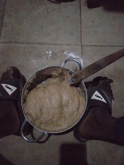 The image captures a top-down view of a food preparation scene taking place indoors, likely in dim lighting. At the center is a large, circular metal pot, possibly aluminum, filled with a thick, brownish-beige, paste-like or dough-like substance. This substance has a slightly uneven surface, suggesting it has been recently stirred or is in the process of being prepared. A large, rustic-looking wooden stick or paddle with a broad, flat end is plunged into the substance within the pot, indicating active stirring or pounding.
The pot rests on a tiled floor, which is light grey or beige with visible dark grout lines forming a grid pattern. Scattered white powdery specks are visible on the tiles around the pot, suggesting that a dry ingredient, possibly flour or starch, has been spilled during the preparation process.
On either side of the pot, a pair of human feet are visible, clad in dark, open-toed sandals or slides. The skin tone of the feet appears dark. The sandals feature a distinct white triangular logo on the strap, with the word "Landry" visible on the left sandal. The feet are positioned as if a person is standing or sitting directly over the pot while engaged in the cooking activity. The overall lighting is dim and uneven, suggesting the scene is taking place in the evening, at night, or in a poorly lit indoor environment.