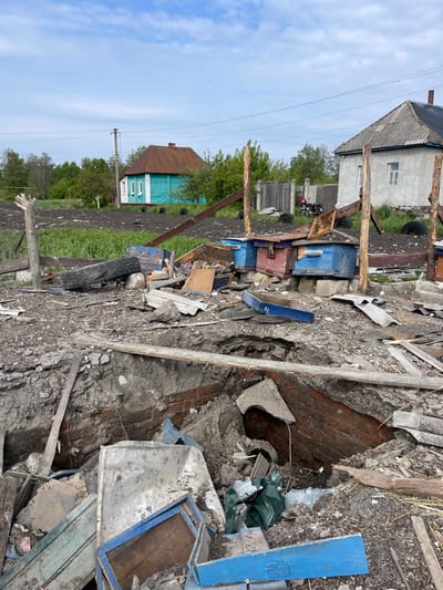 The scene is a residential area in Bilopillia, Ukraine, located at 51.139 latitude, 34.285 longitude. There is debris scattered on the ground, including wood, metal, and brick. A large hole in the ground is visible, which appears to be a damaged basement or cellar. Several wooden beehives are present, some of which have been damaged. There are two small houses in the background, one painted blue and the other white. The environment is overgrown with grass and weeds, and there are trees in the background. The scene appears to be the aftermath of a bomb strike or other violent event.
