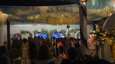 Indoor event with numerous attendees. Overhead decorations feature intricate draping and chandeliers. A stage area with white floral arrangements and geometric shapes is visible. People are gathered in groups, and some are dressed formally. The lighting is soft, creating a warm ambiance. Tables and chairs are arranged for seating. The scene appears to be a wedding reception.