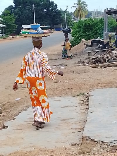 This media file depicts a street scene in Iseyin, Nigeria. The primary subject is a person wearing a vibrant orange and white striped robe with circular yellow patterns, carrying a large tray of food items balanced on their head. The tray holds various bowls filled with what appears to be produce or prepared food, suggesting a vendor or someone transporting goods for sale or consumption.

In the background, other individuals are visible. To the right, a child in a yellow shirt and patterned skirt stands near some building materials and firewood, possibly a construction or market area. Further back, another person in a patterned garment is seen near what might be cooking facilities. The scene is set outdoors on a dirt road with a paved walkway in the foreground. Lush green trees and vegetation frame the background, indicating a tropical or subtropical environment. Building structures, including what looks like a partial house and some scaffolding, suggest ongoing development or a mixed residential and commercial area.

The time of day appears to be late afternoon or early evening, judging by the soft lighting and the overcast sky. The weather seems calm and possibly humid, typical for the region. The overall atmosphere is one of everyday life and commerce in a Nigerian community. There is no visible text in the image.