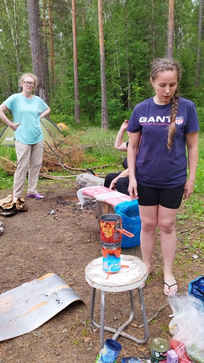 In Orekhovo, Russia, amidst a dense forest, a group of individuals are engaged in an outdoor activity. The foreground features a portable camping stove set atop a rustic, white stool. Next to the stool, a blue plastic bag and various food items, including cans and what appears to be fruit, are visible. In the background, a woman with glasses stands with her hands on her hips, wearing a light blue t-shirt and beige pants. To her right, another woman, her hair in a braid, is dressed in a dark blue t-shirt and black shorts. Two tents are set up behind them. The ground is covered with pine needles and dirt, and fallen branches are scattered around the campsite, indicating a natural, wooded environment. The overall scene suggests a recreational gathering or camping trip in a forest setting.